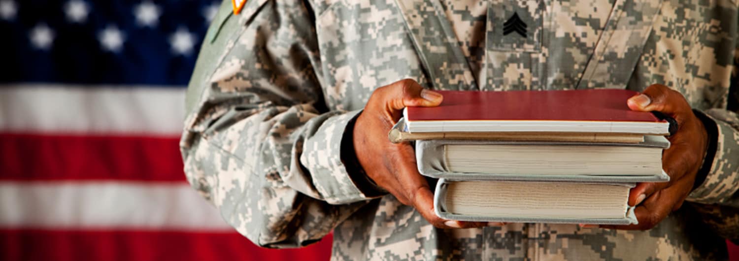An enlisted military member standing in front of an American flag holding a Bible, journal, and M.Div. textbooks.