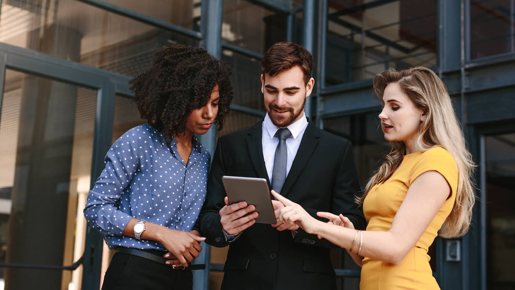 Two women and one man, dressed in business attire, standing outside a modern office building and looking at a tablet together.
