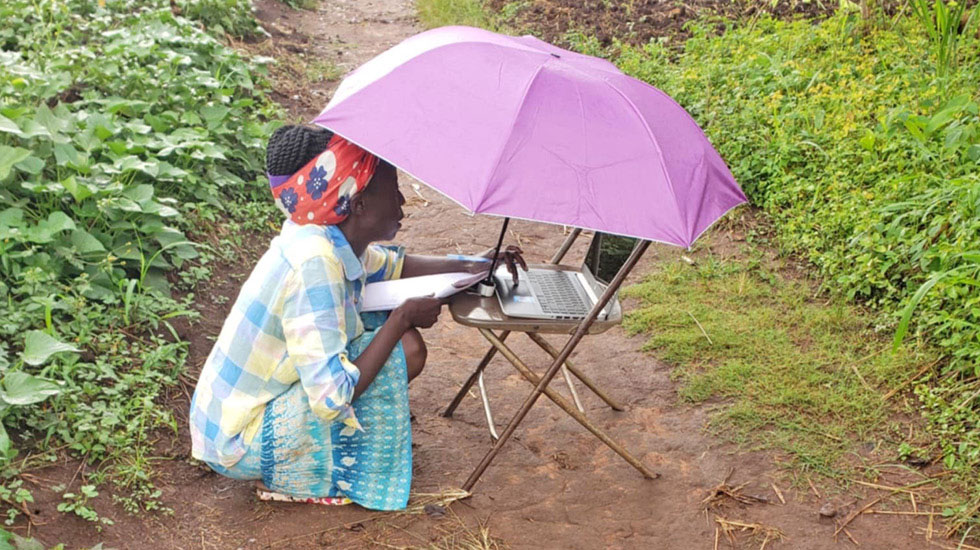 Regent University online student Prudence Nakaweesa on her laptop in the rain outside her village.