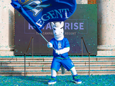 Rex the Royal, Regent University's white horse mascot, waving a large Regent flag at a campus event surrounded by blue and green confetti.