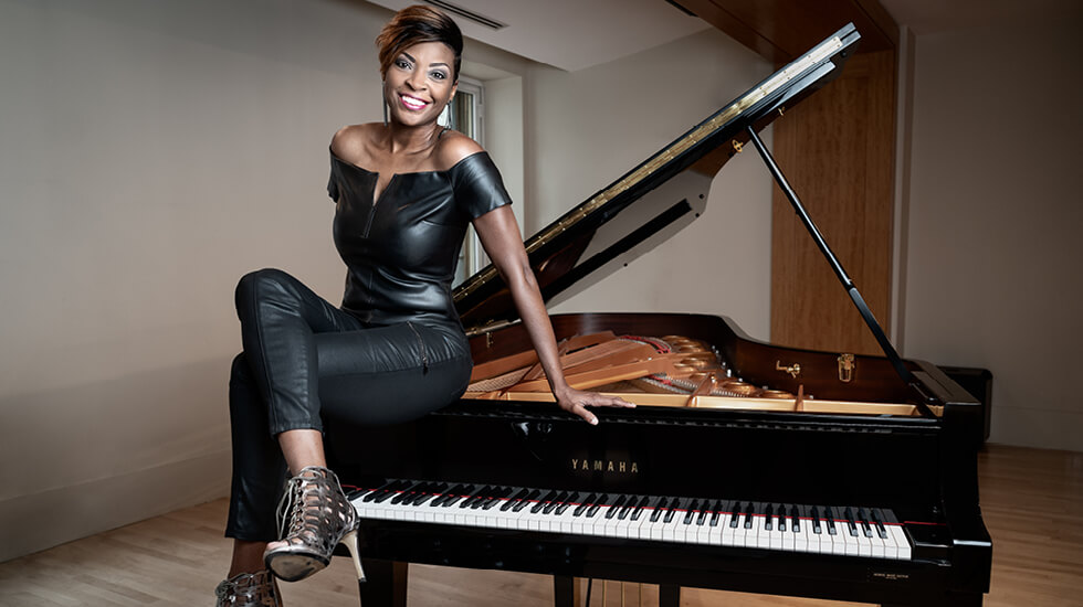 Jade Simmons, dressed in black, smiling while seated on a grand piano in a performance hall