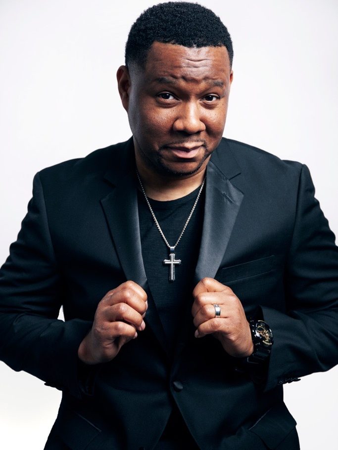Mr. Talkbox  in black suit wearing a cross necklace.