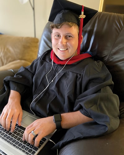 Preston Braswell in graduation regalia sitting with laptop, Class of 2020