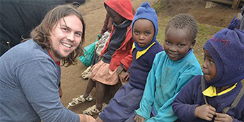 Kenton Lee, Regent University alumnus and founder of The Shoe That Grows, smiles beside children in Kenya wearing school uniforms and jackets.