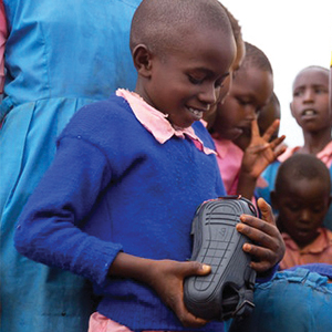 A young child in Kenya smiles while holding a pair of The Shoe That Grows, an adjustable shoe designed to expand in size as the child grows.