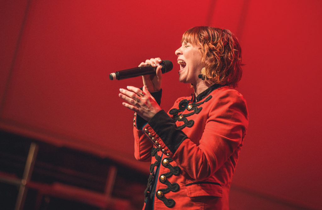 Worship leader in red jacket singing passionately into microphone against red backdrop