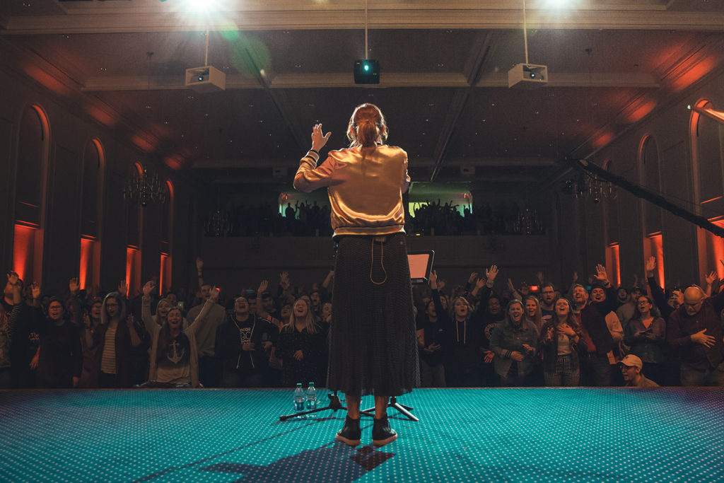 Worship leader on stage facing congregation with raised hands in chapel
